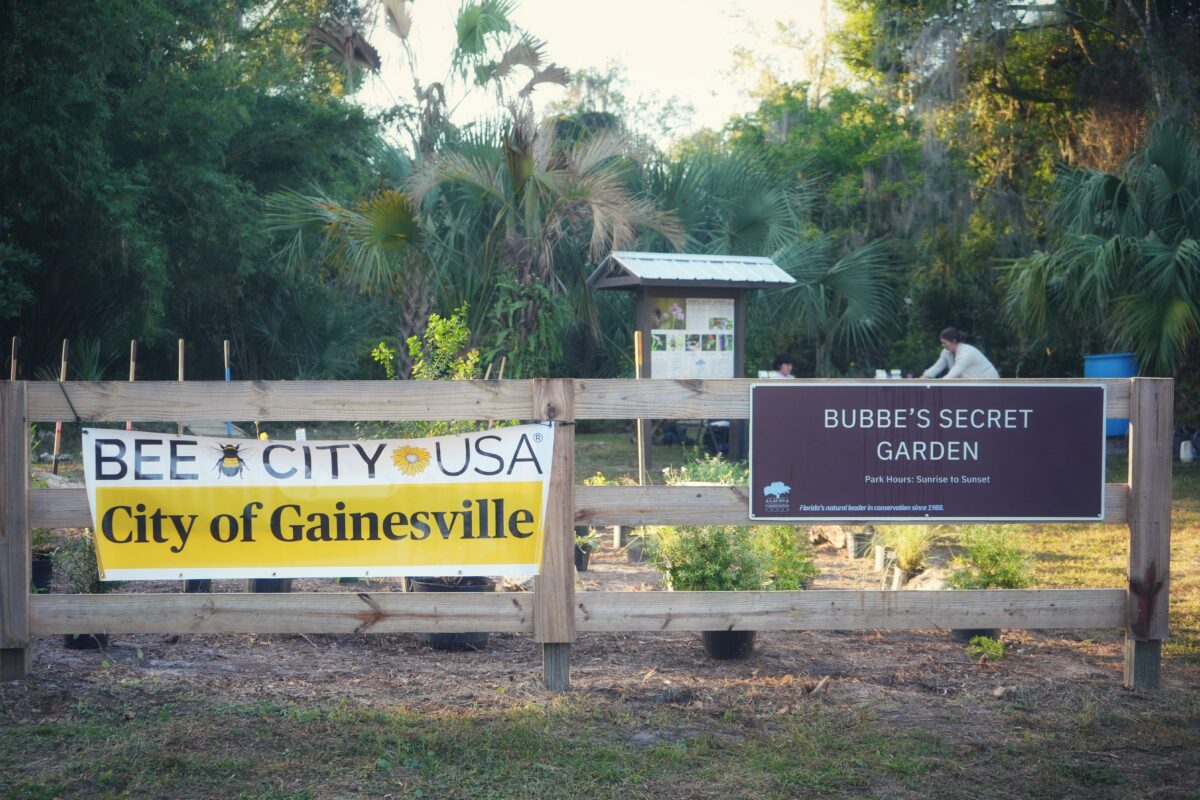 Community garden with signs reading Bee City USA and Bubbe's Secret Garden in Gainesville. People work in the background, surrounded by trees.