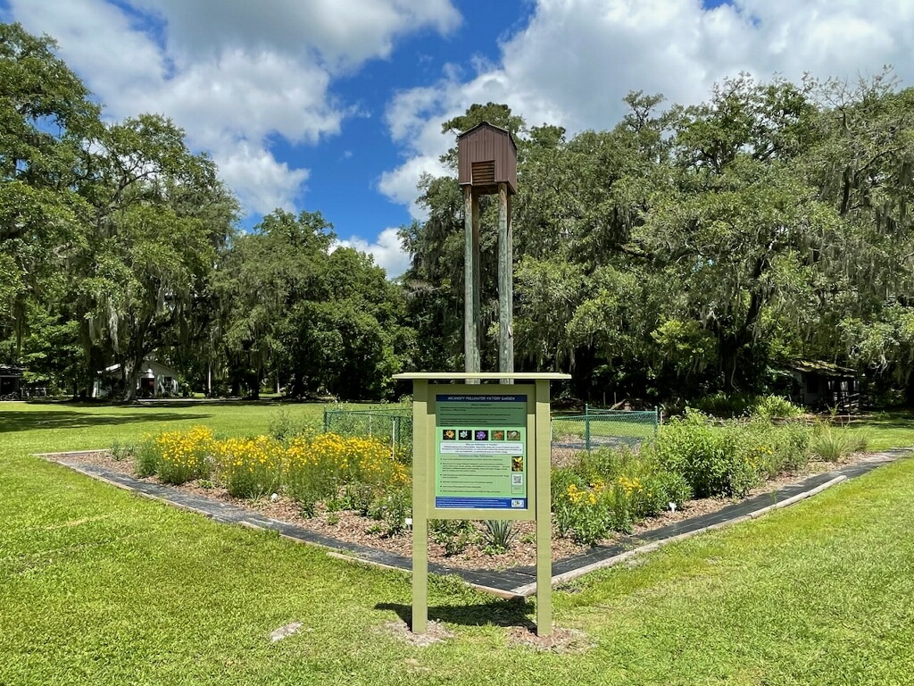 A wooden bat house sits atop a pole near an informational sign in front of a garden with yellow flowers surrounded by trees and a blue sky with clouds.