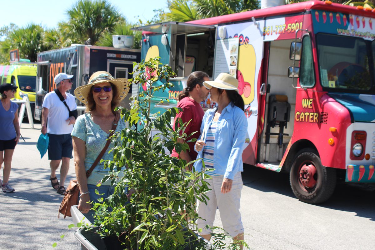 A woman in a sun hat stands near food trucks, surrounded by people and potted plants, on a sunny day.